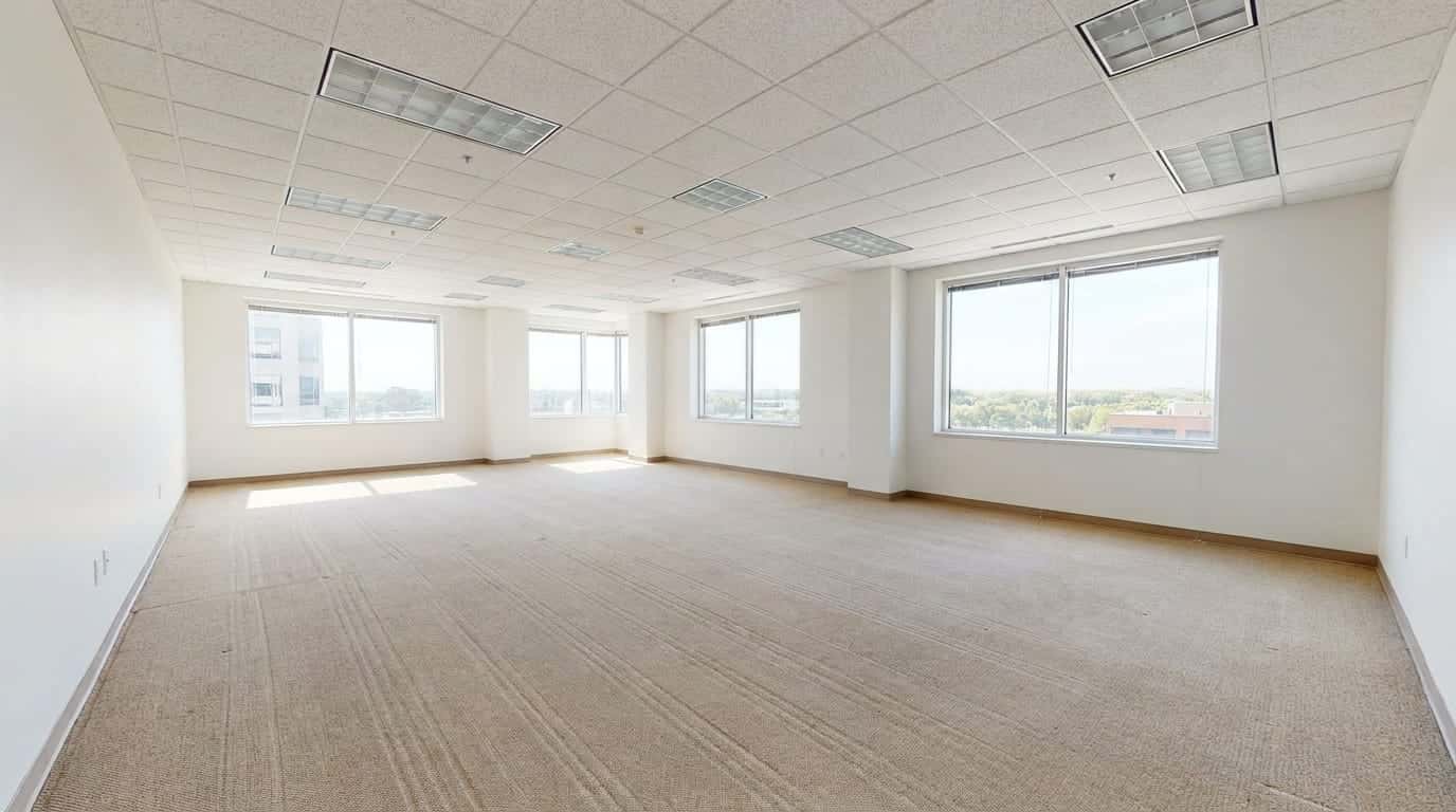 An empty office suite with beige carpet showing fresh vacuum lines, white walls, a suspended ceiling and large windows letting in daylight