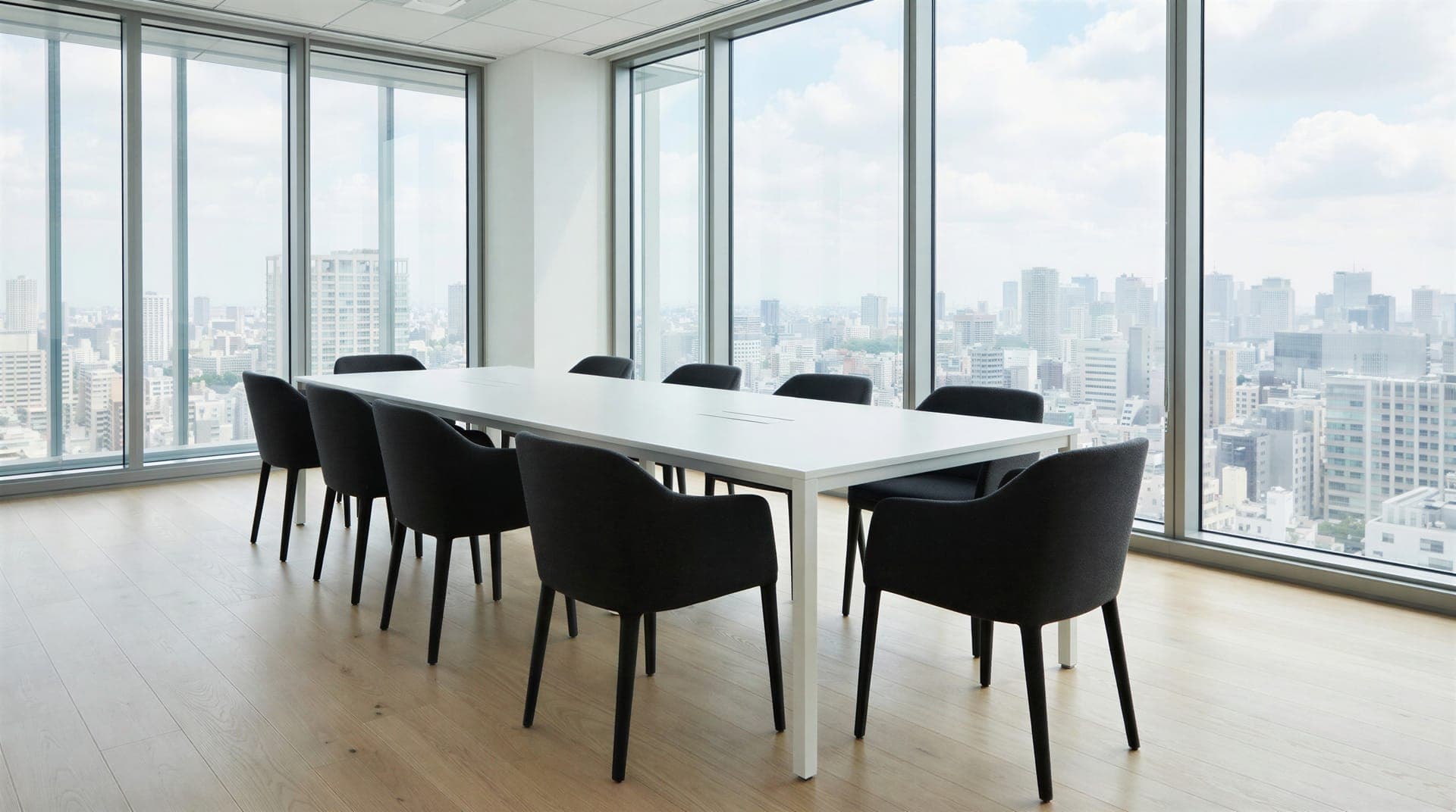 A meeting room with a long white table, dark upholstered chairs, a pale wood floor and floor-to-ceiling windows looking over a city